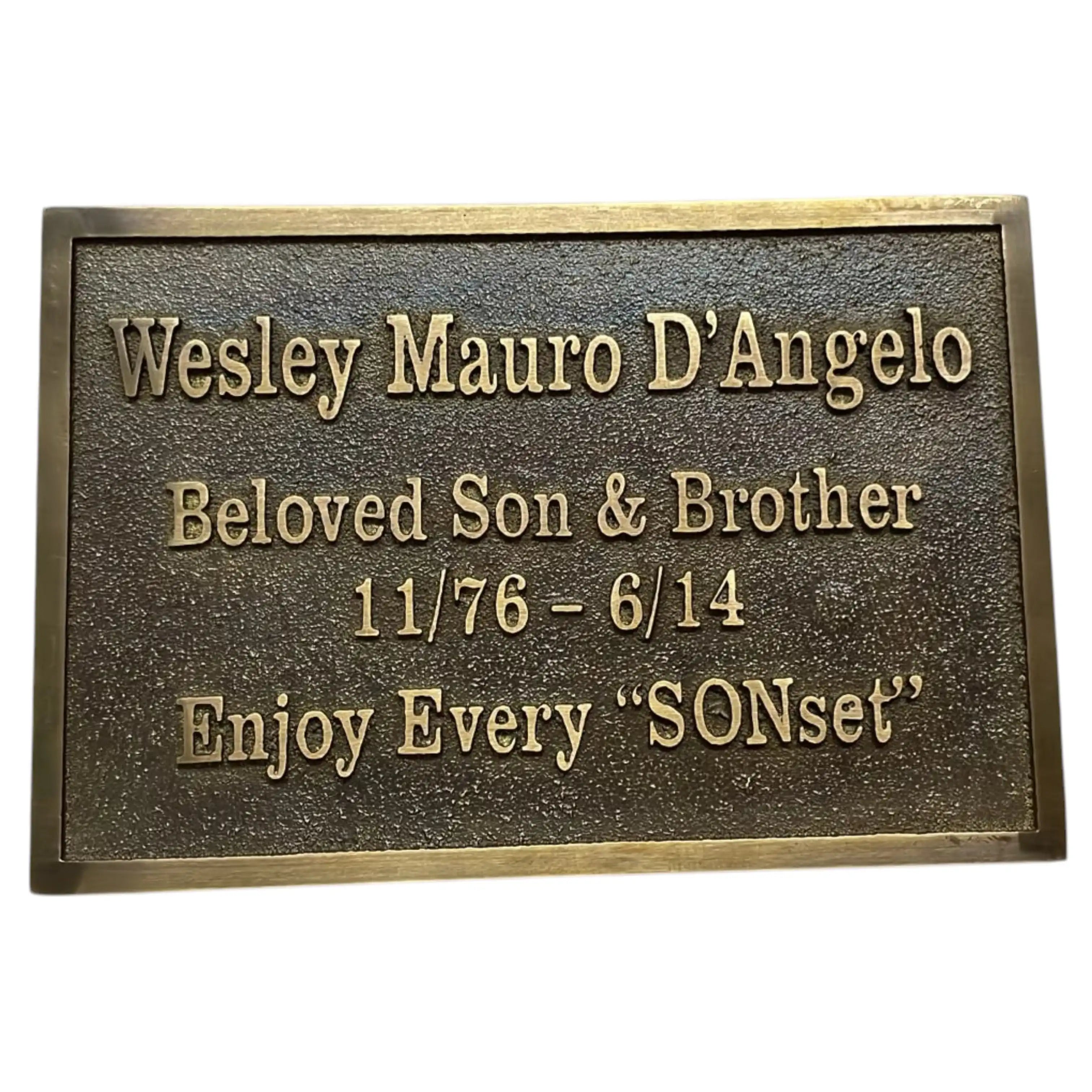 A rectangular bronze memorial plaque with raised lettering that reads 'Wesley Mauro D'Angelo, Beloved Son & Brother, 11/76 - 6/14, Enjoy Every "SONset"' on a plain white background.