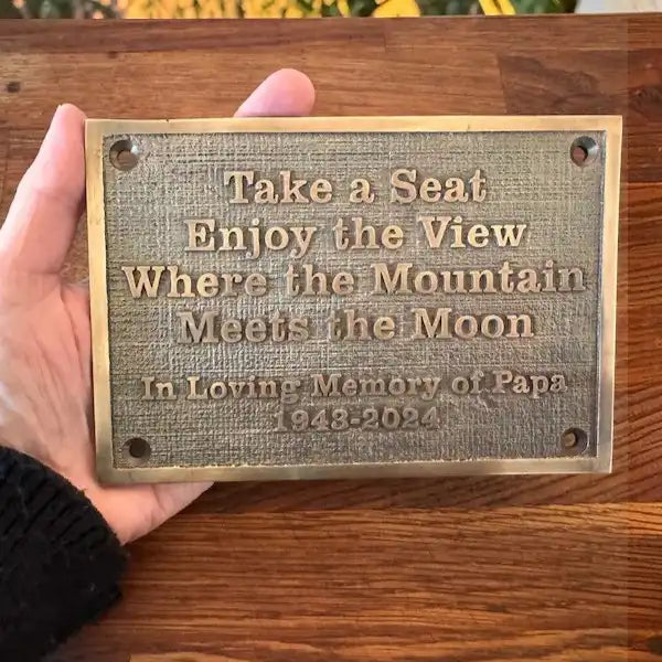 Hand holding a brass plaque with engraved text on a wooden surface