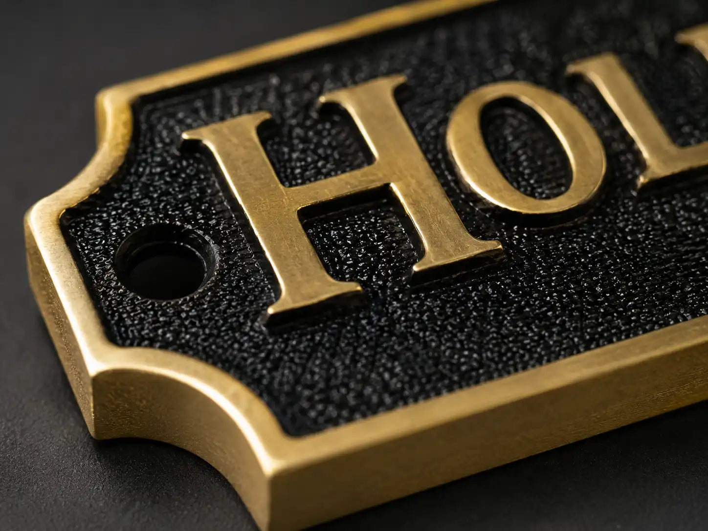 Close-up of a metal plaque or sign with raised gold lettering reading "HOL" on a textured black background, photographed on a dark surface.
