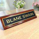 A personalized wooden desk nameplate with a polished reddish-brown wood base, a black textured center panel, and gold lettering reading 'BLANE SWAIN', shown on a light wood tabletop with blurred flowers in the background.